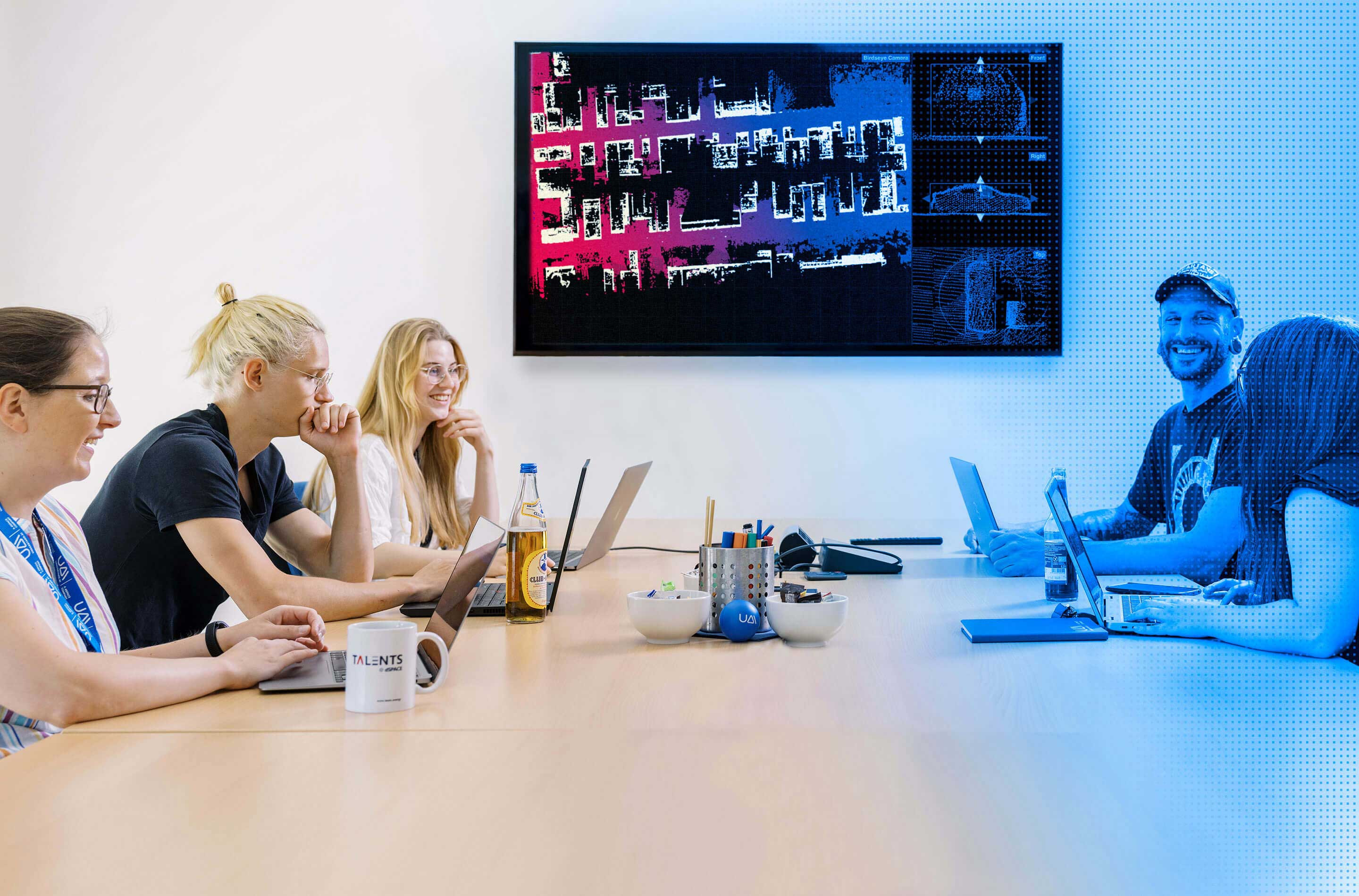 Team members seated in an office with laptops and stationery on a table, next to a screen displaying Ground Truth graphics on the wall