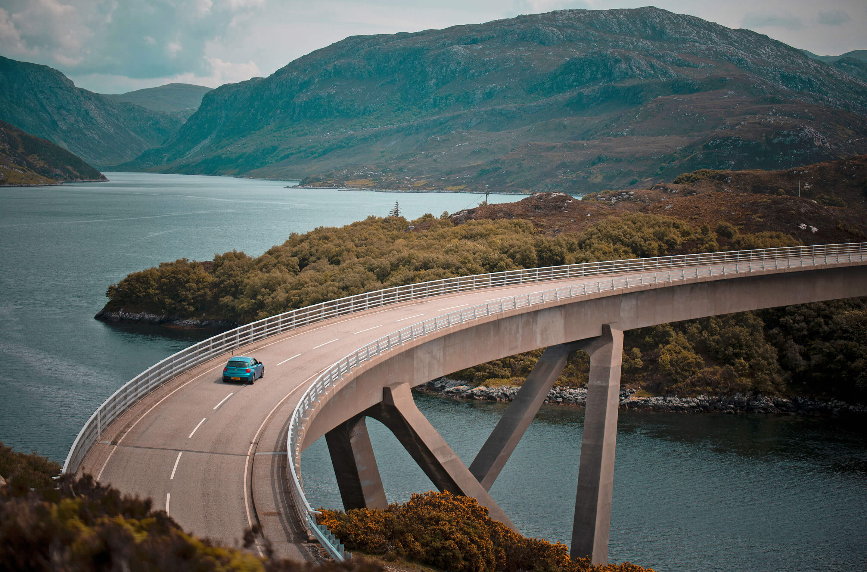 Scenic view of a curving bridge over calm waters with a solitary vehicle, framed by rugged mountains and coastline